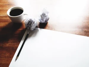 Leeres Blatt Papier, Stift, zerknüllte Notizen und eine Tasse Kaffee – Symbolbild für Trauerkarte schreiben.